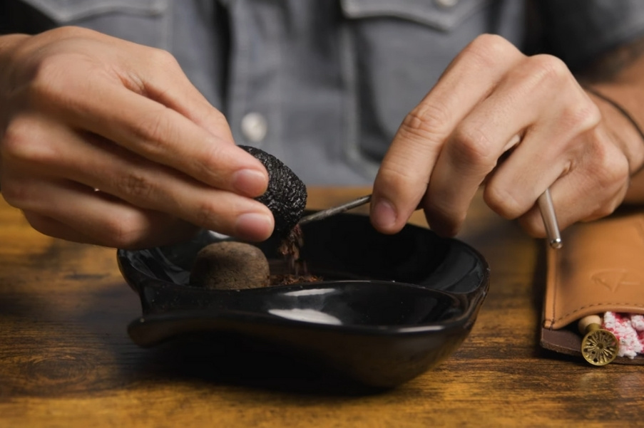 Hands using a pipe tool to clean tobacco residue from a smoking pipe over an ashtray, demonstrating post-smoke maintenance.