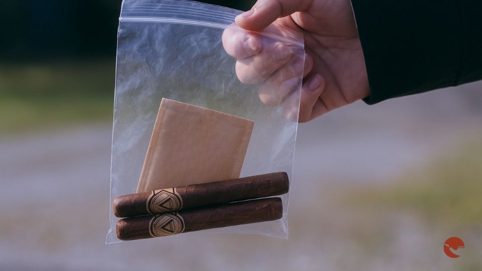 Mano che regge un sacchetto di plastica trasparente con sigari e una bustina umidificante Boveda in un ambiente esterno.